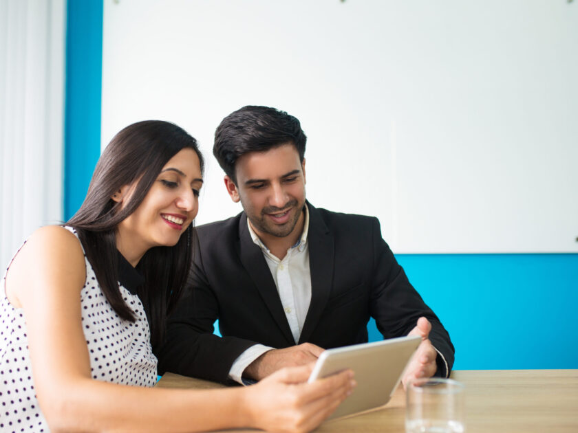 Portrait of happy young colleagues using digital tablet. Young Caucasian businessman and businesswoman sitting at table and watching data on pc tablet. Wireless technology in business