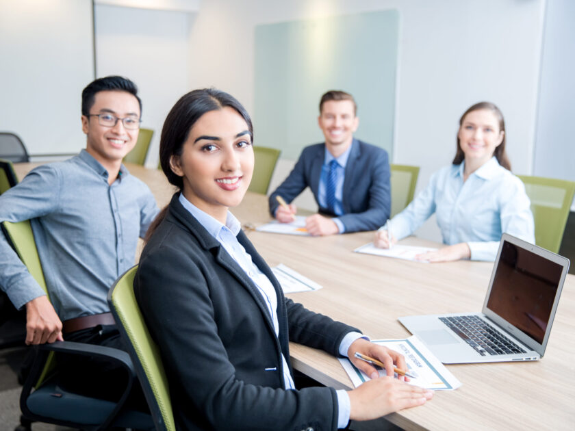 Four smiling middle-aged multi-ethnic business people turning to camera, working and discussing ideas while sitting at big table in conference room. Side view.